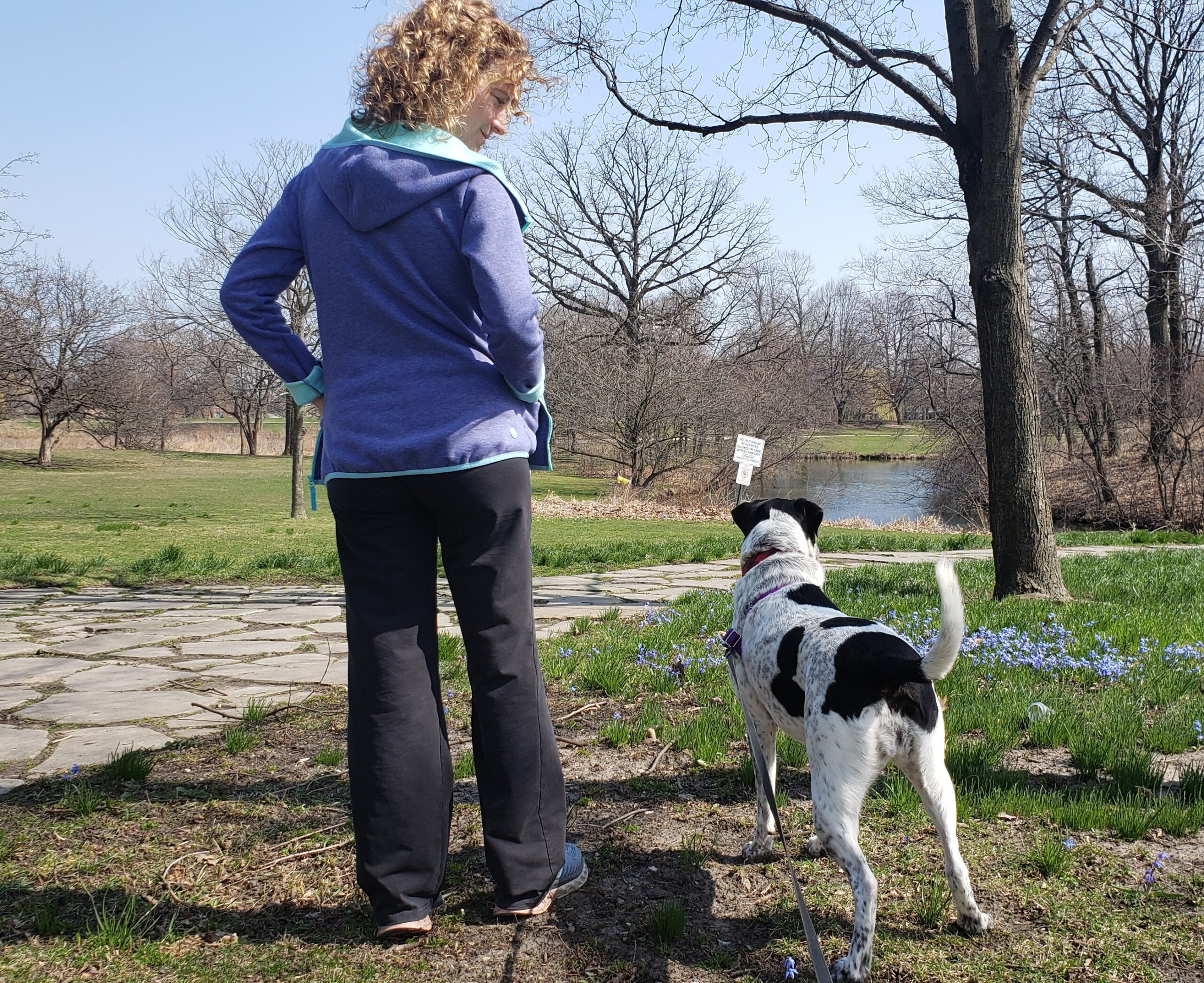 Woman standing beside a black and white dog on a leash in a grassy park near a pond.